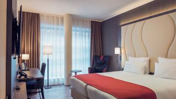 A hotel room with a bed, a desk, a chair, a lamp, and a TV.