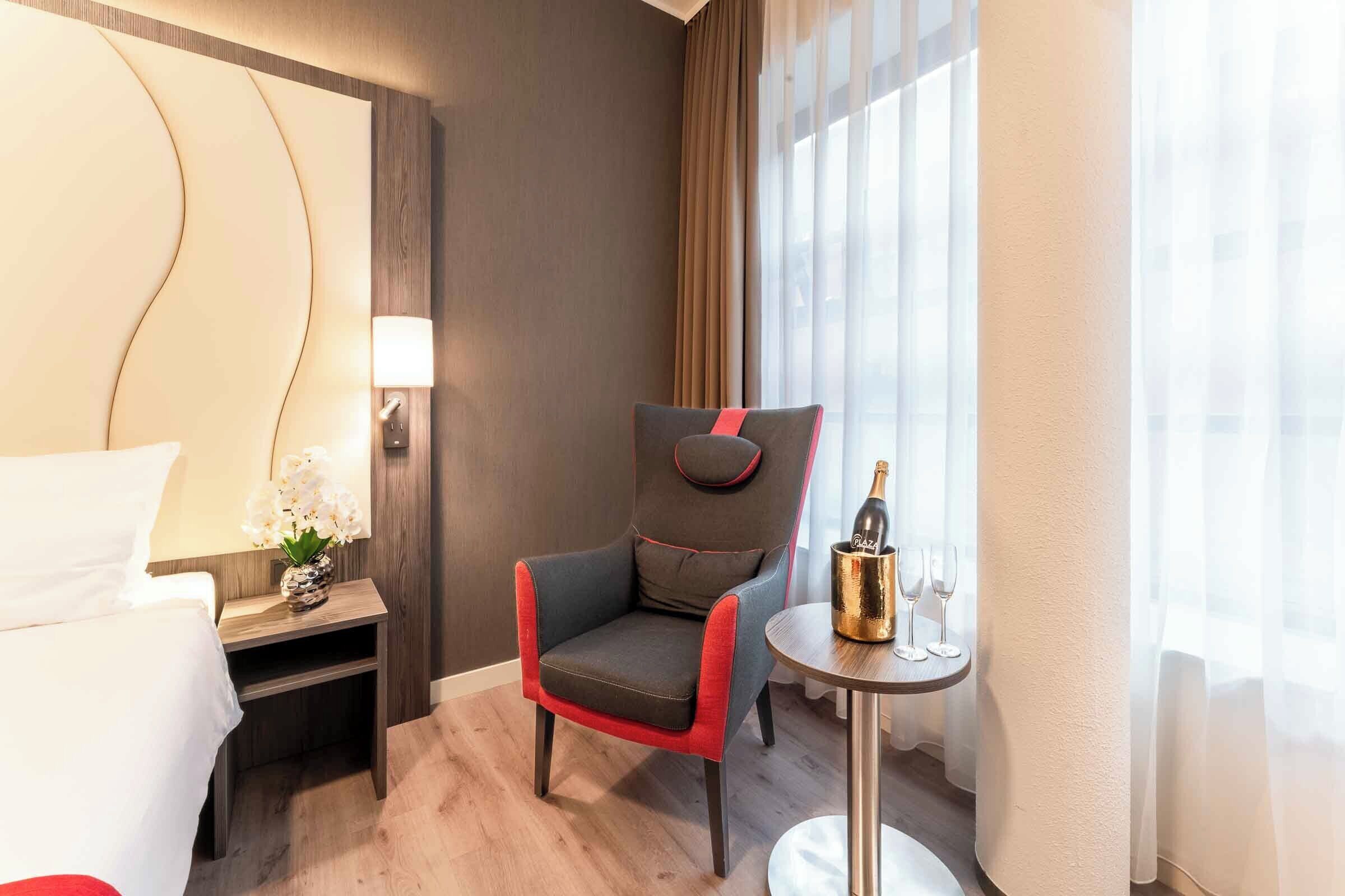 A hotel room with a bed, a chair, a small table with a bottle and glasses, and a window with curtains.
