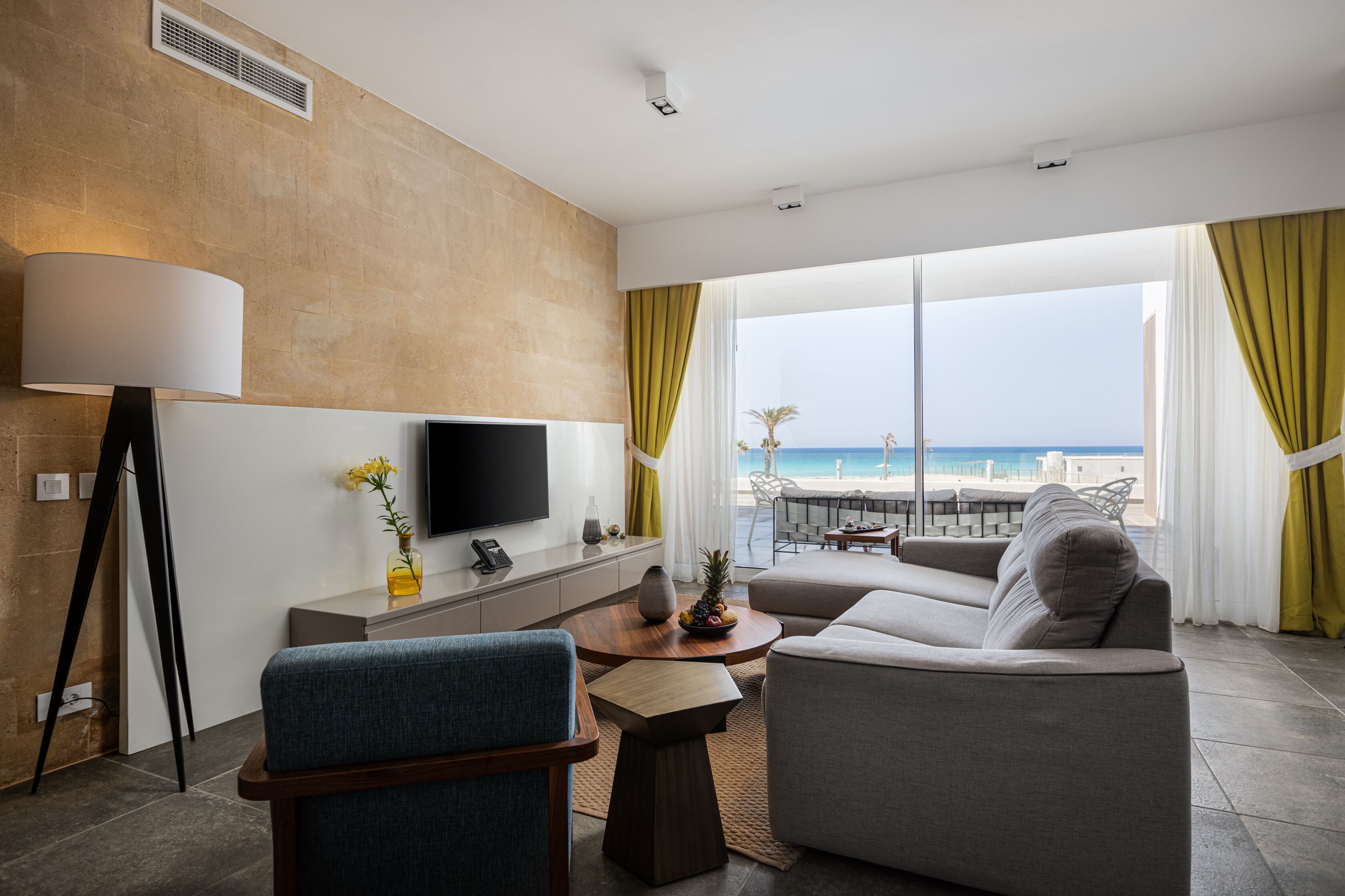 A modern living room with a sofa, armchair, and coffee table, a flat-screen TV, and a view of the beach.