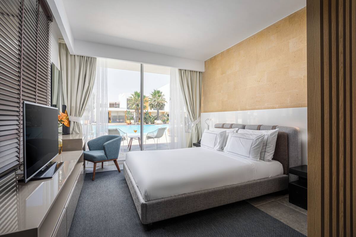 A modern hotel room with a large bed, a TV, a desk, and a view of a pool and palm trees.