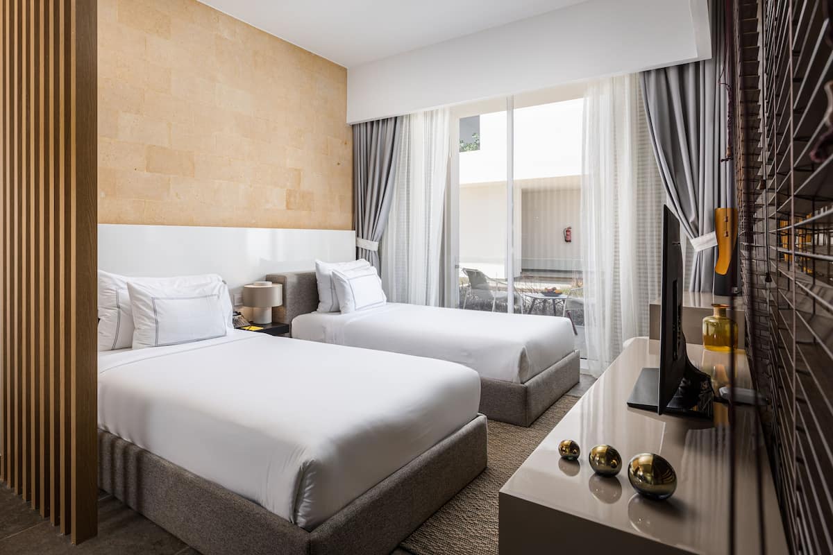 A hotel room with two beds, a desk with a TV, and a view of the outdoors.