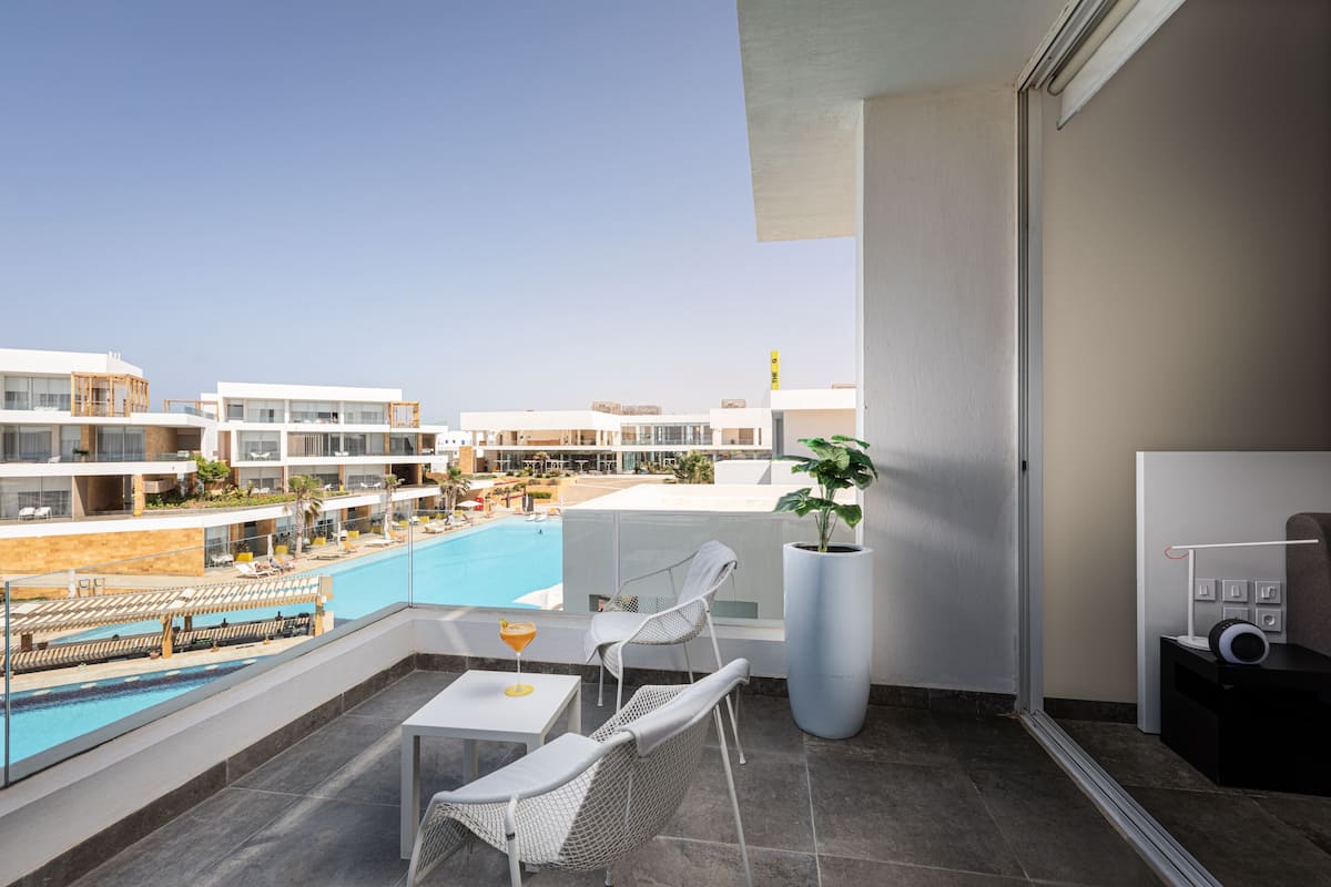 A balcony with a pool view, white chairs, a table with a drink, and a large potted plant.
