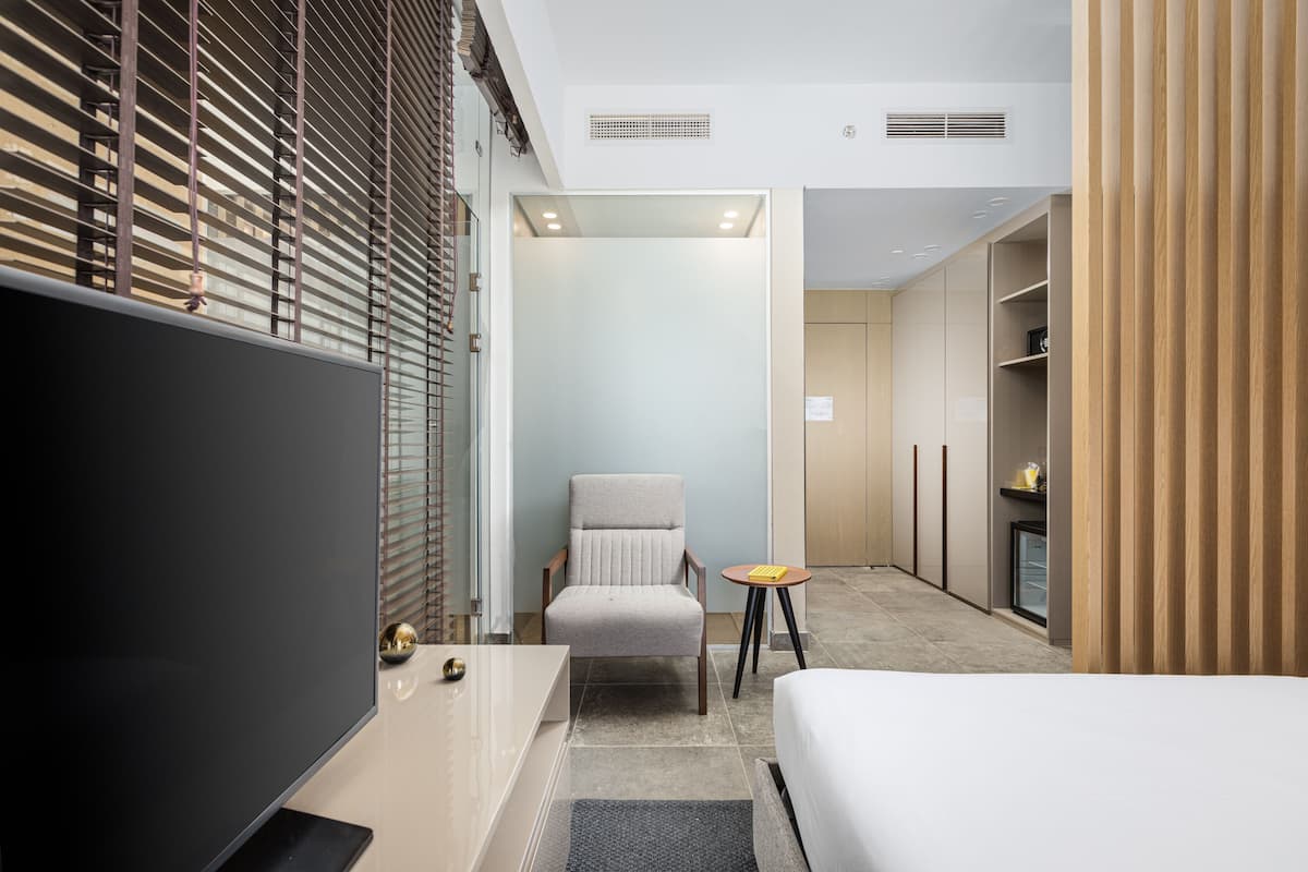 A modern hotel room with a large bed, a seating area with a chair and a small table, a television, and a wardrobe.