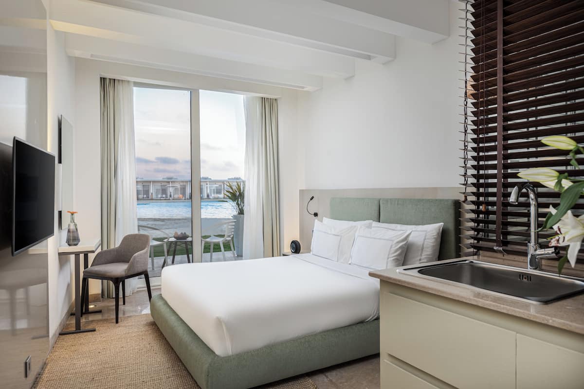 A hotel room with a bed, a desk, a TV, a window with curtains, a view of the water, and a sink.