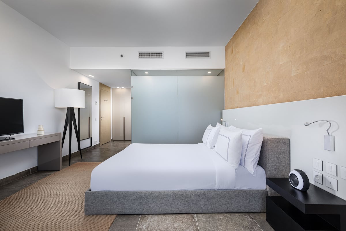 A modern hotel room with a large bed, a desk with a lamp, a flat-screen TV, and a wall-mounted speaker.