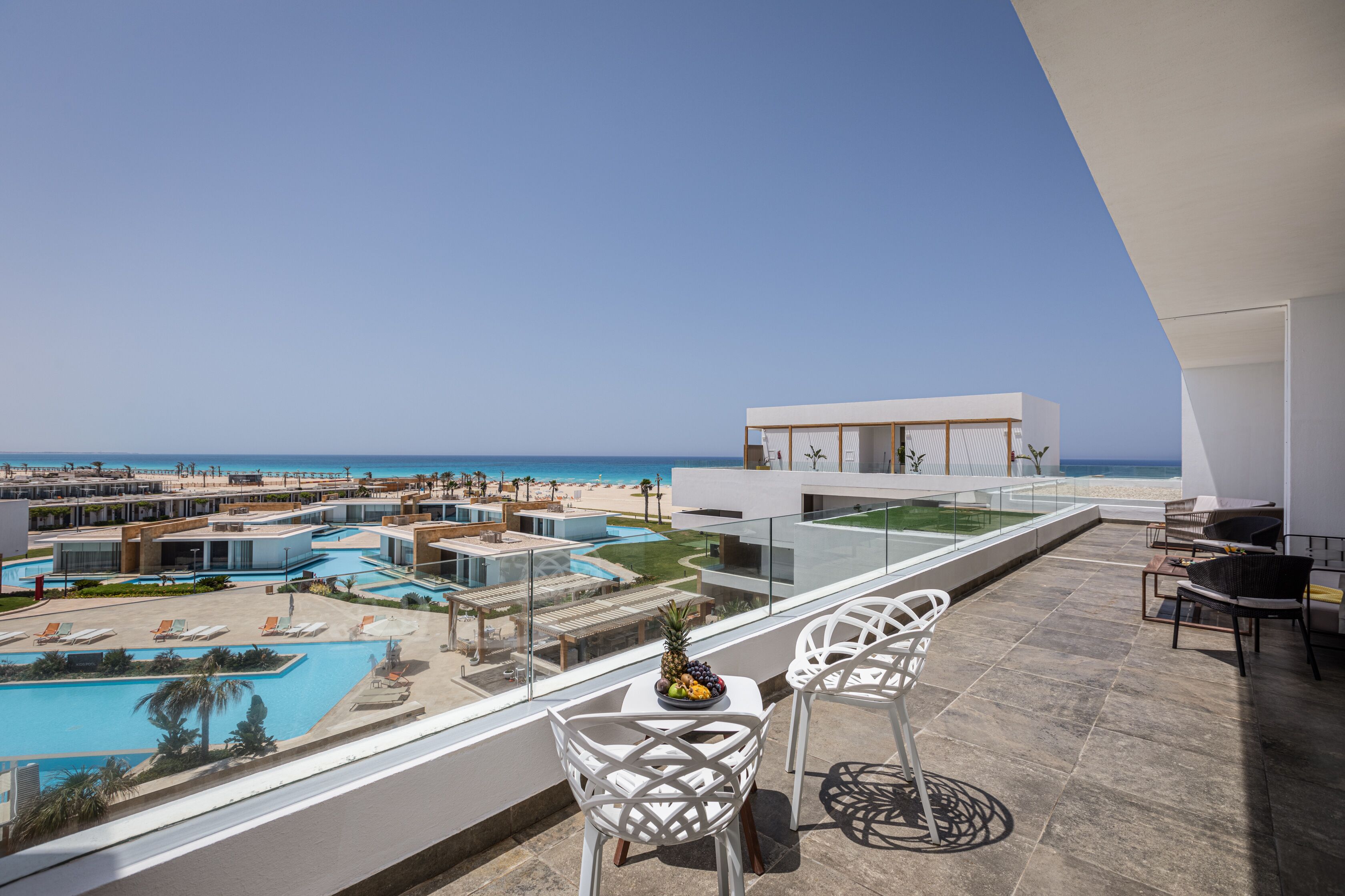 Un balcon avec des chaises blanches, une table avec des fruits, et une vue sur une piscine et une plage.
