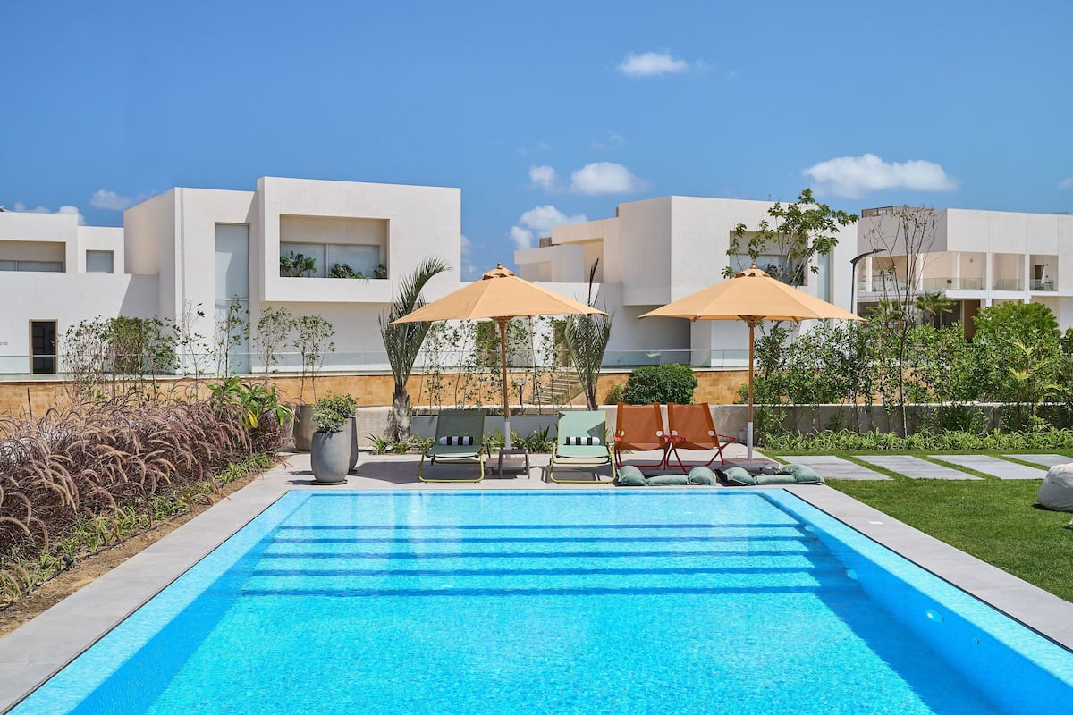 A swimming pool with lounge chairs and umbrellas, surrounded by modern buildings and greenery.