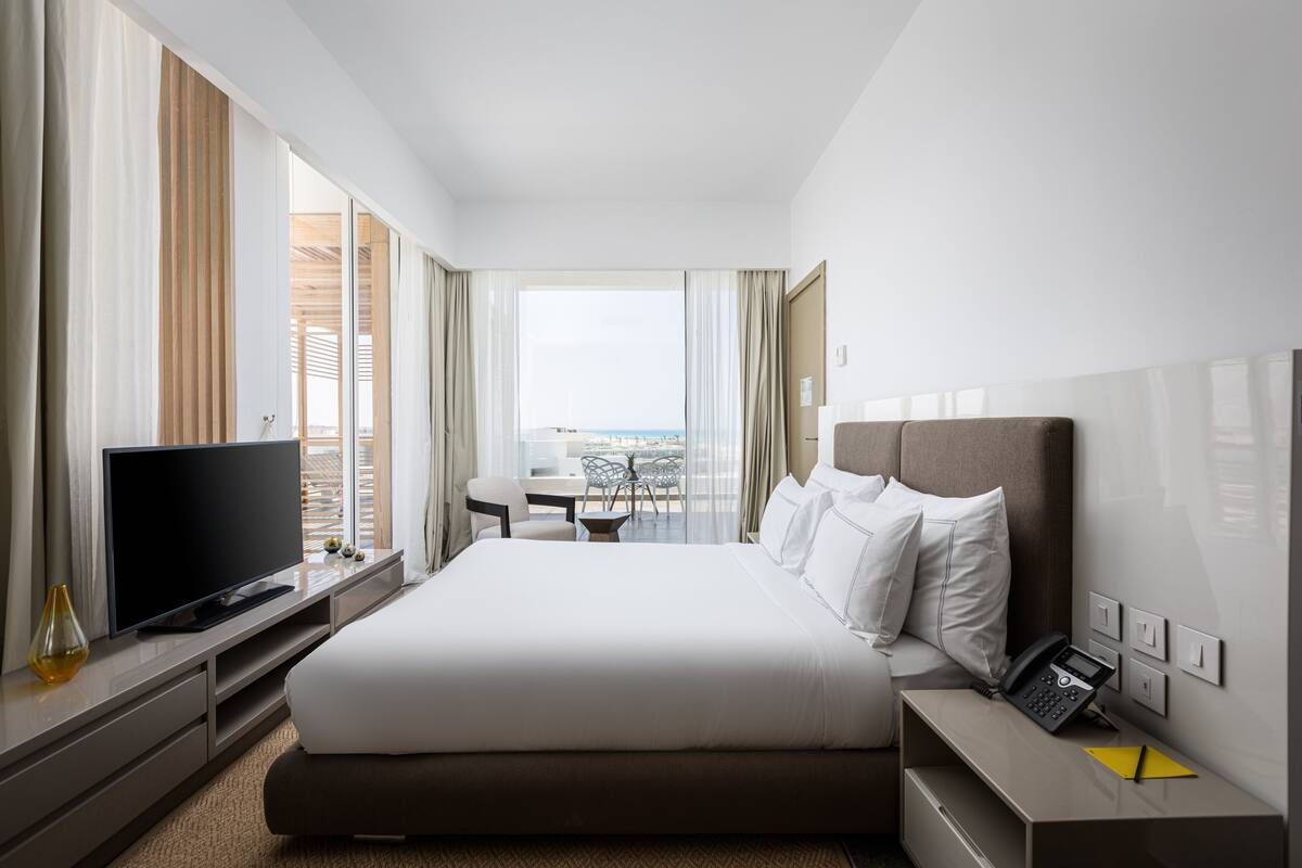 A modern hotel room with a large bed, a flat-screen TV, a desk with a chair, and a balcony with a view.