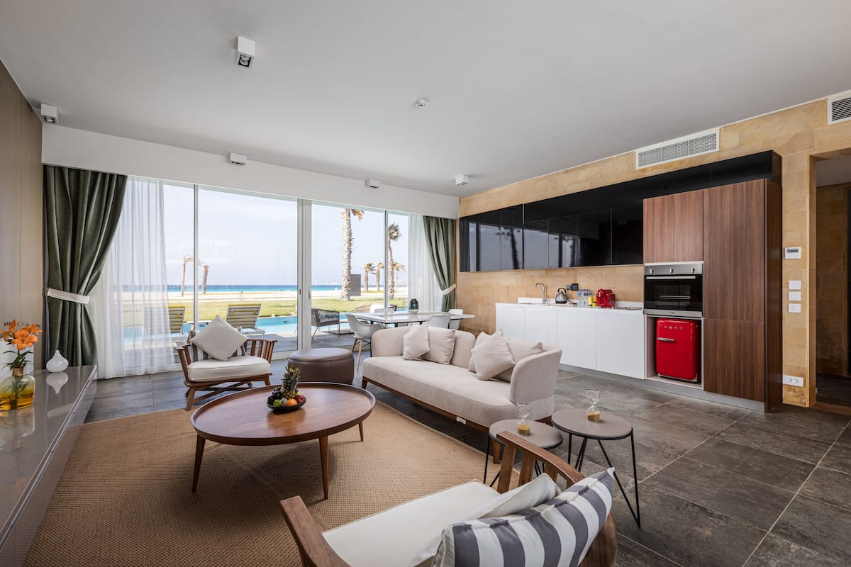 A modern living room with a sofa, coffee table, and chairs, a kitchen with a red fridge, and a large window with a view of the beach.
