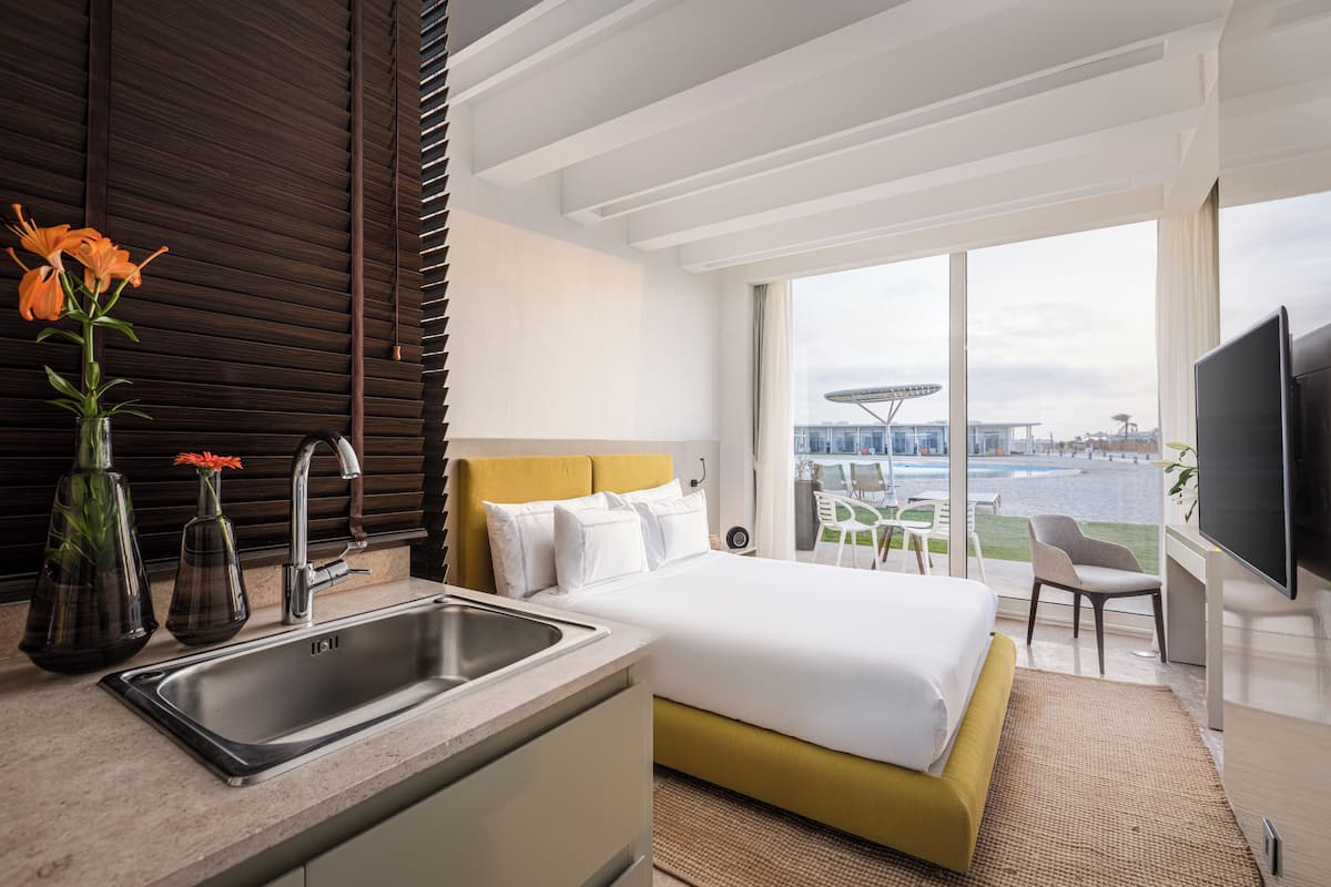 A modern hotel room with a large bed, a sink, and a view of a cityscape through large windows.