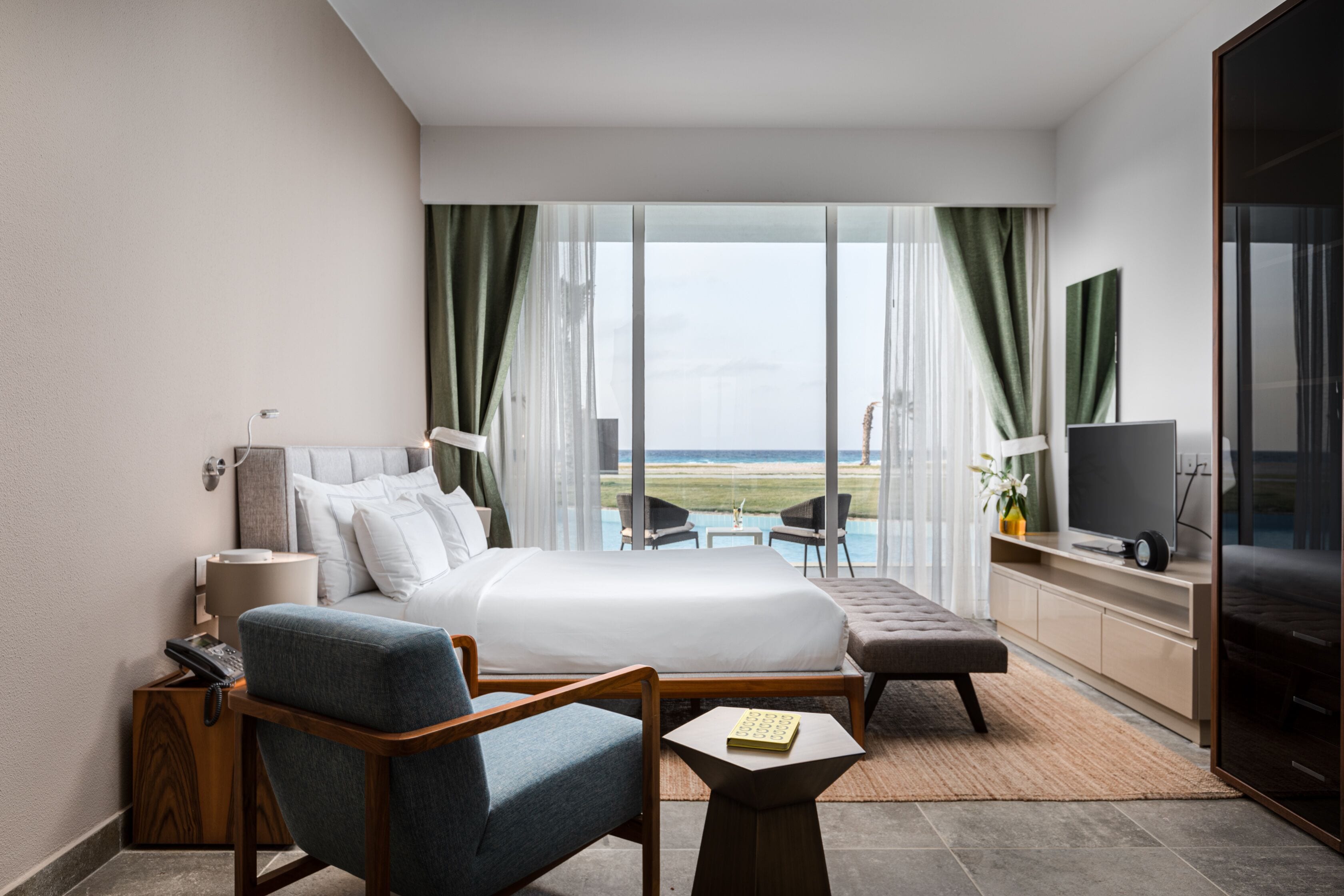 Habitación de hotel moderna con una cama grande, zona de estar con una silla y una mesita, televisión y vistas a la playa a través de amplios ventanales.