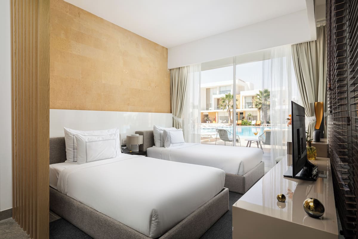 A hotel room with two beds, a TV, a desk, and a view of a pool and buildings.