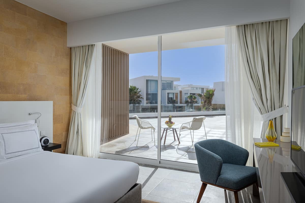 A hotel room with a bed, a desk, a chair, and a balcony with a view of modern buildings.