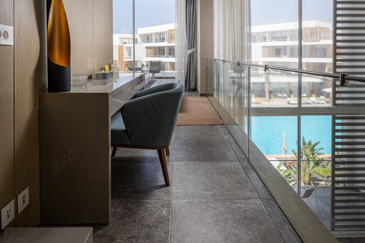 A modern hotel room with a balcony, a dining area, and a view of the pool and buildings.