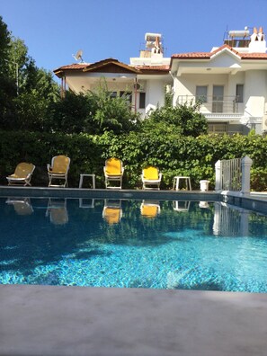 A swimming pool with clear blue water, surrounded by greenery and a white building with a tiled roof in the background. - Ustun Apart (Marmaris)