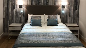 A modern bedroom with a wooden wall panel design, a large bed with a patterned duvet, two bedside tables, and two wall-mounted reading lamps.