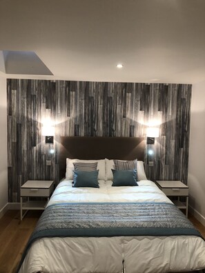A modern bedroom with a wooden wall panel design, a large bed with a patterned duvet, two bedside tables, and two wall-mounted reading lamps. - Hamilton Arms (Midhurst)