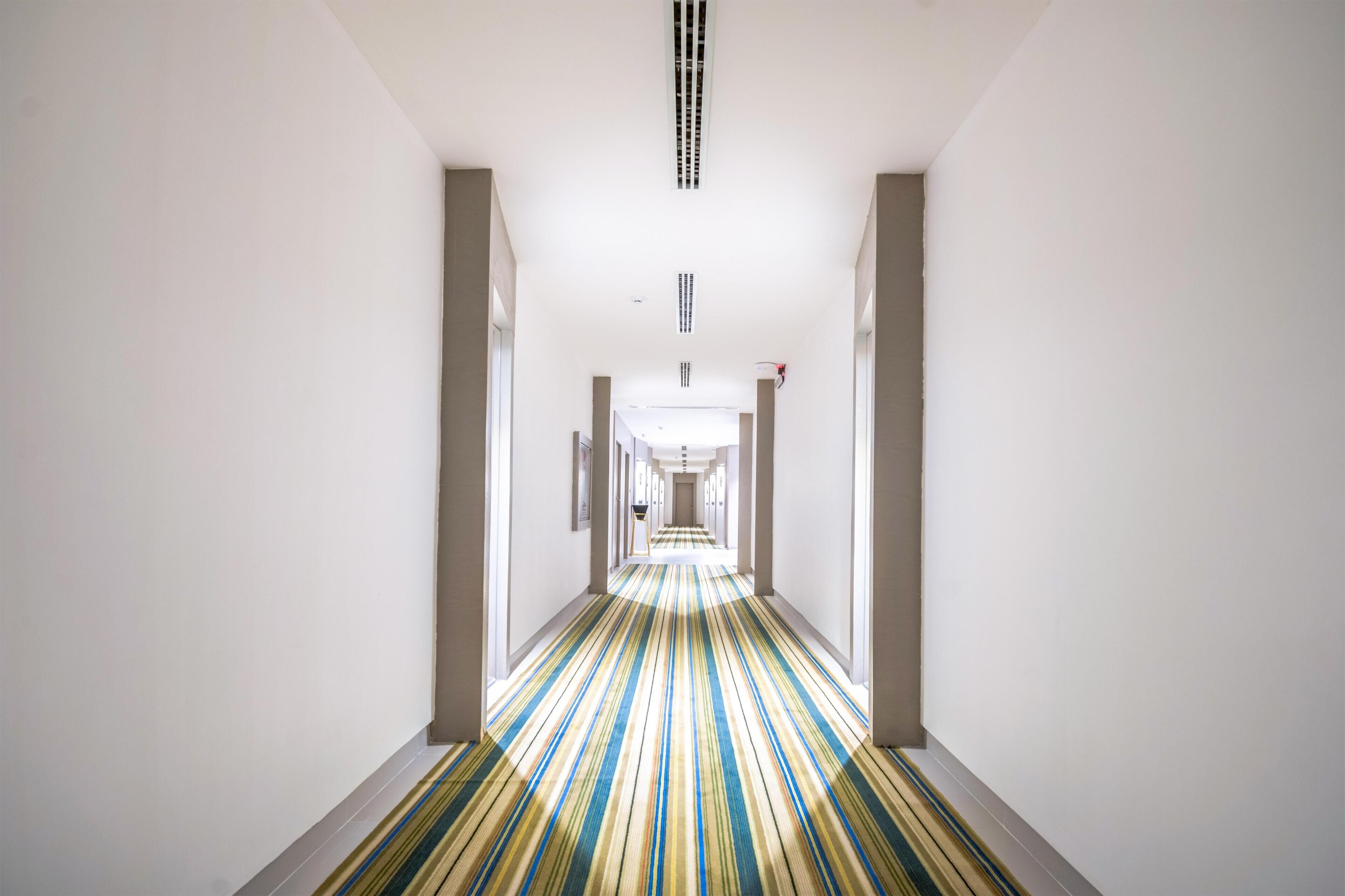A long, narrow hotel hallway with a striped carpet, white walls, and a ceiling with a vent.