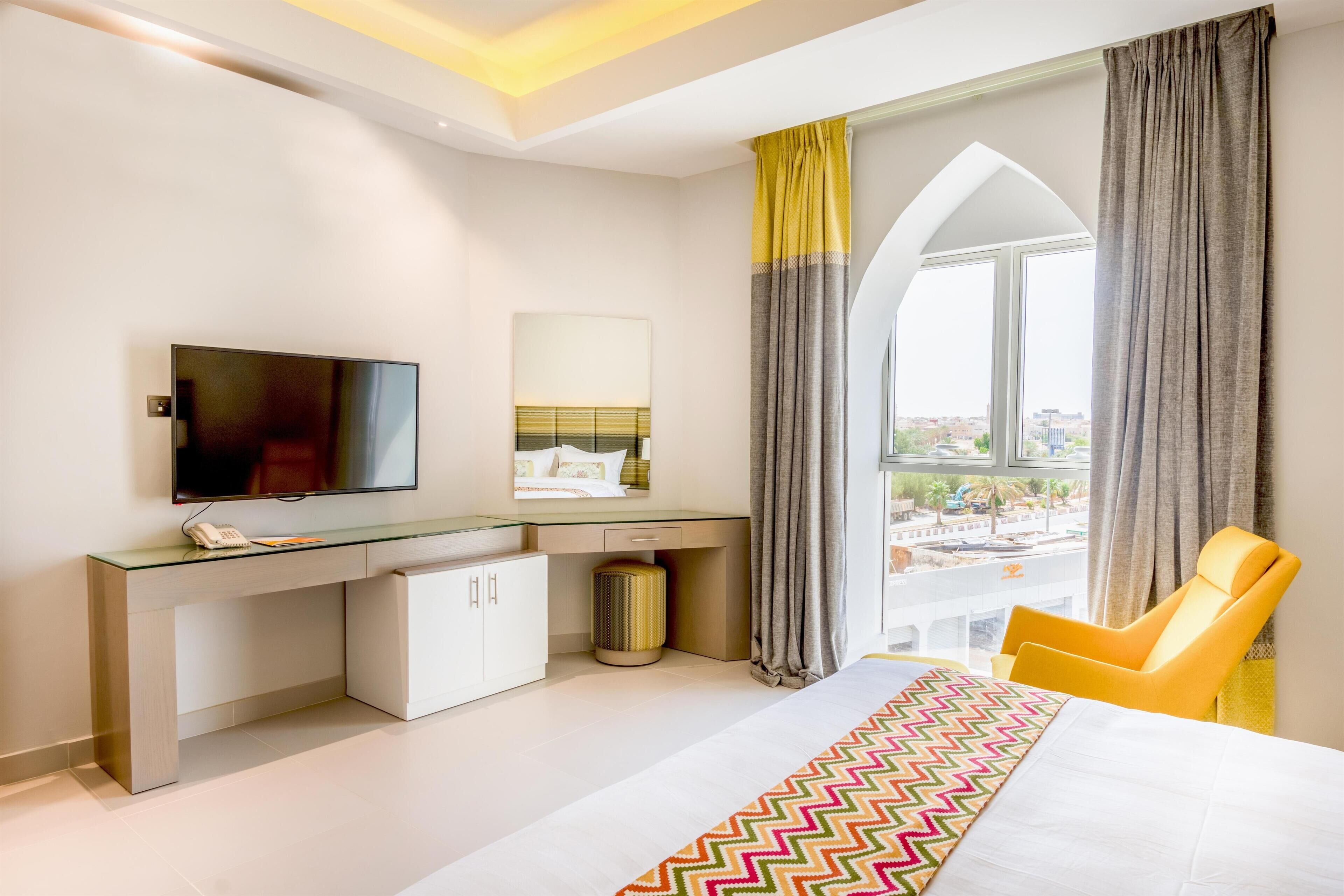Une chambre d’hôtel moderne dotée d’un grand lit, d’un bureau avec un téléviseur à écran plat, d’une chaise et offrant une vue sur la ville grâce à une grande fenêtre.