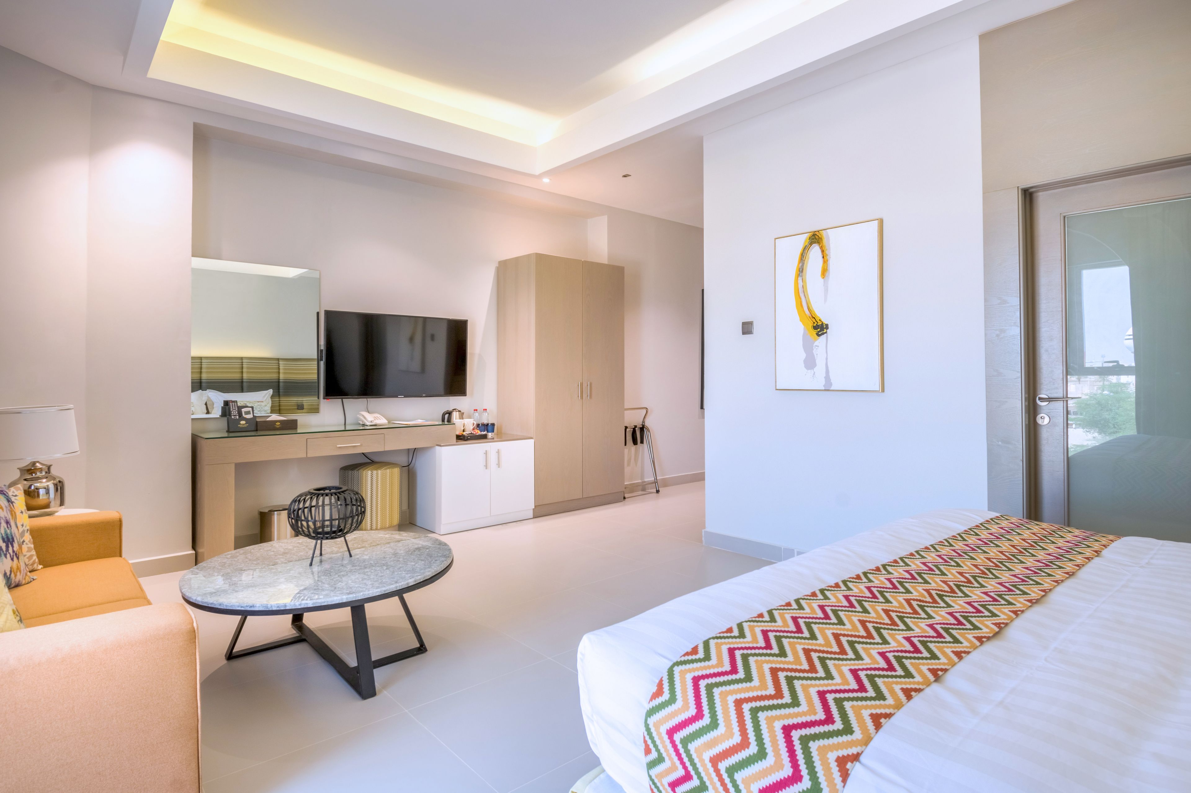 Una habitación de hotel moderna con cama, una pequeña mesa redonda, un sofá, televisor, minibar y un ventanal grande.