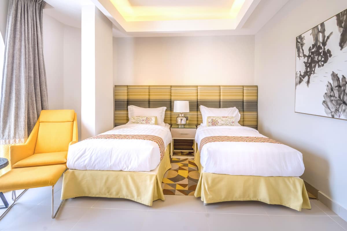 Ein Hotelzimmer mit zwei Betten, einem gelben Sessel, einem Nachttisch mit Lampe und einem großen abstrakten Gemälde an der Wand.