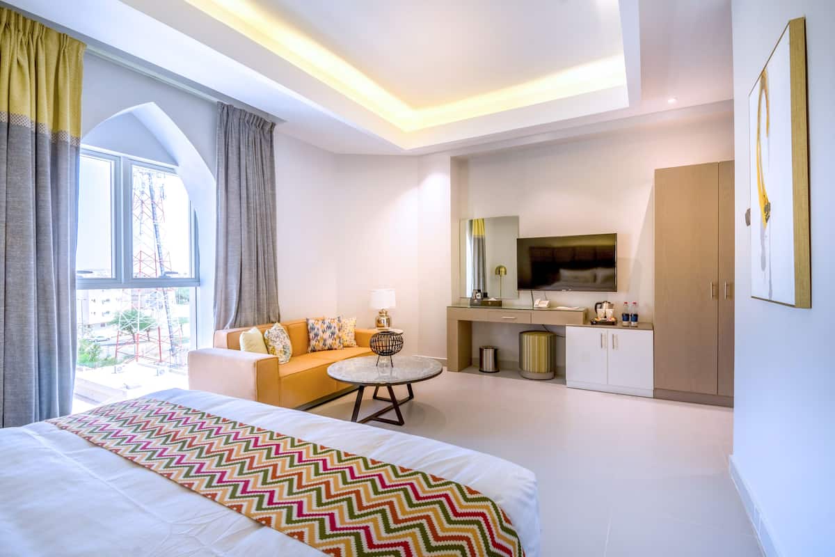 Ein modernes Hotelzimmer mit einem großen Bett, einer Couch, einem kleinen runden Tisch, einem Fernseher und einer Küchenzeile.