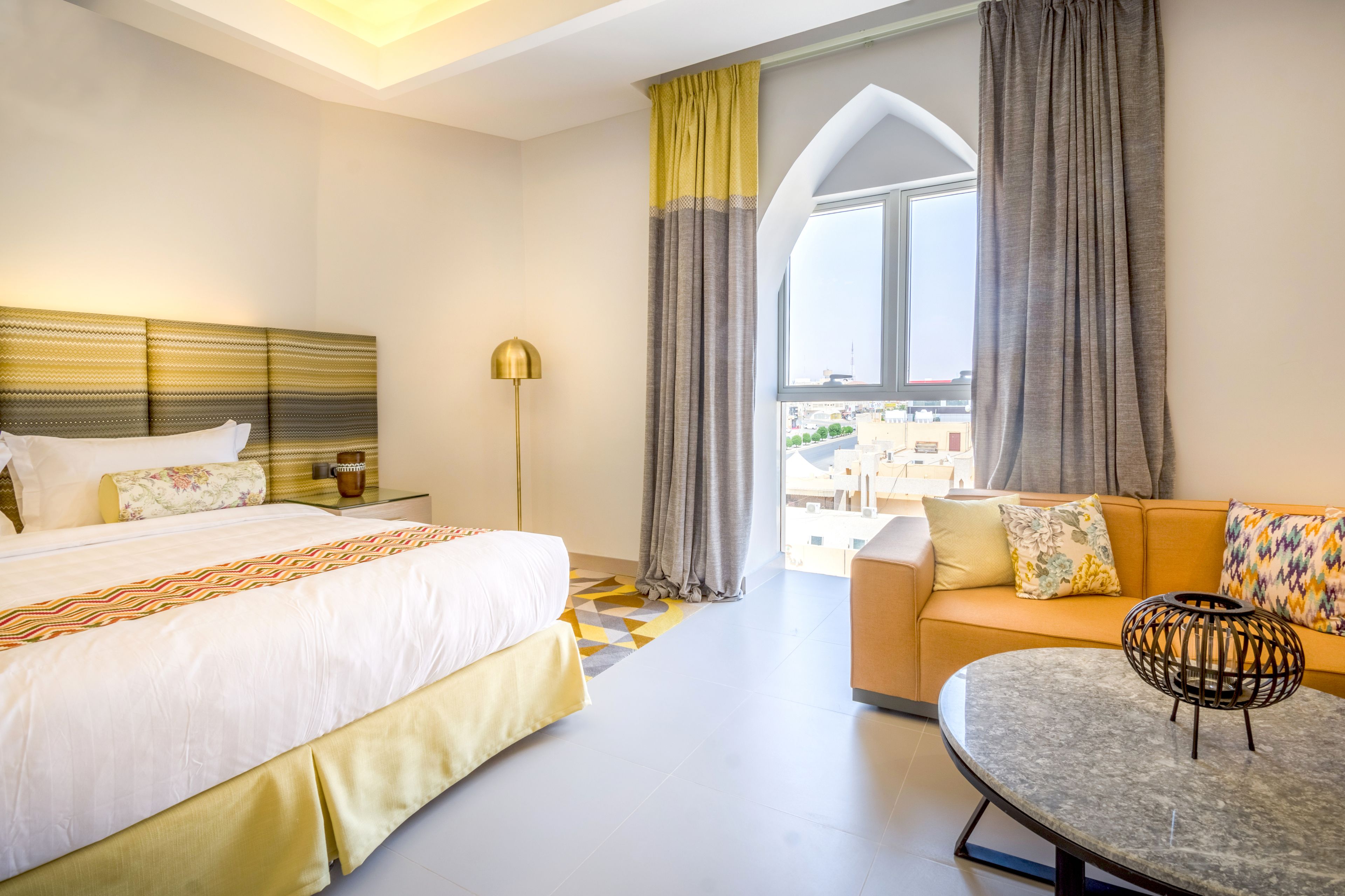 A hotel room with a bed, a sofa, a round table, and a view of the city through a window.
