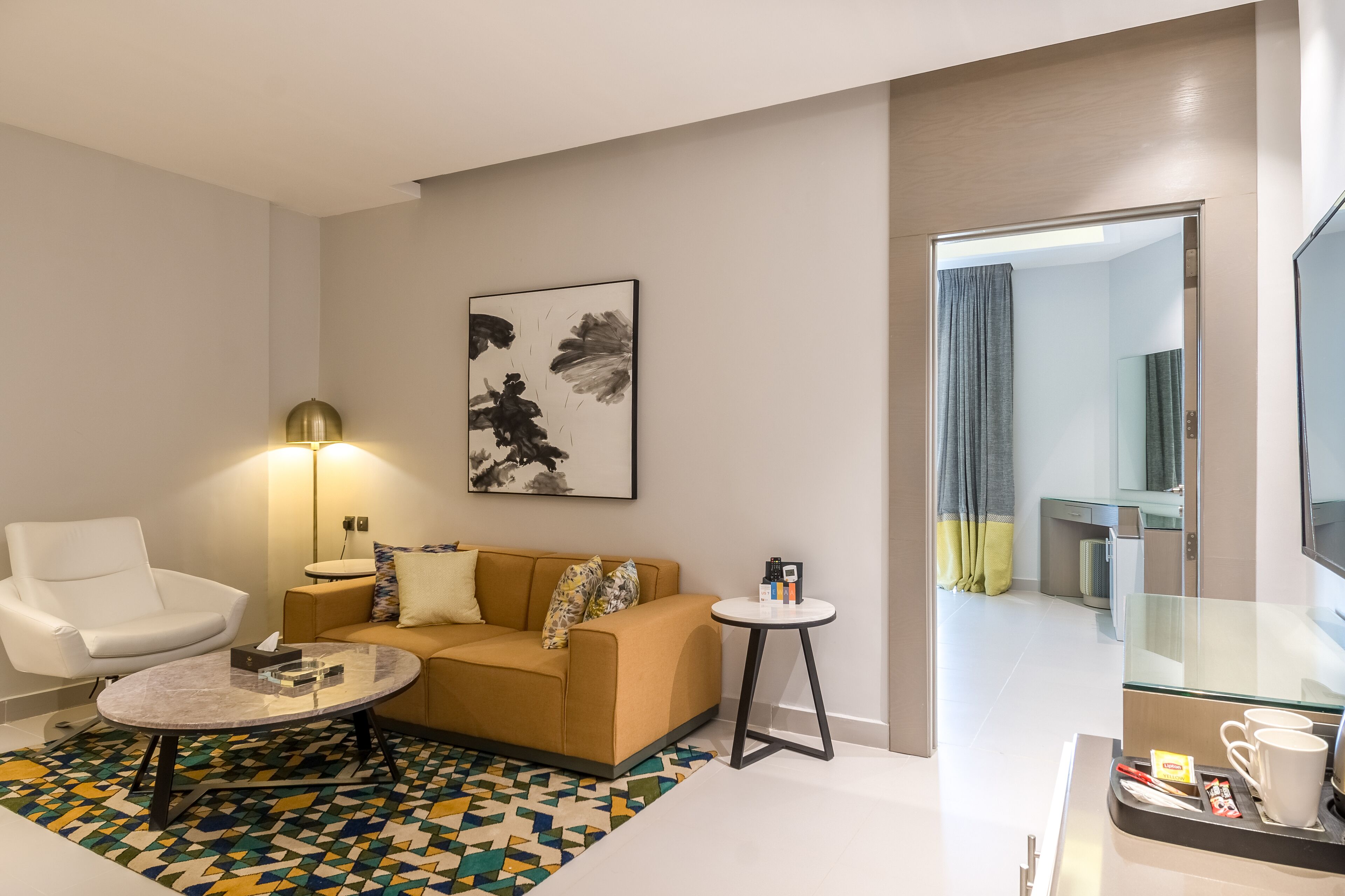 Une chambre d’hôtel moderne avec un fauteuil blanc, un canapé beige, une table basse ronde, une petite table d’appoint, une grande peinture abstraite, une table basse en verre, un tapis à motifs et une cloison en verre agrémentée d’un miroir.