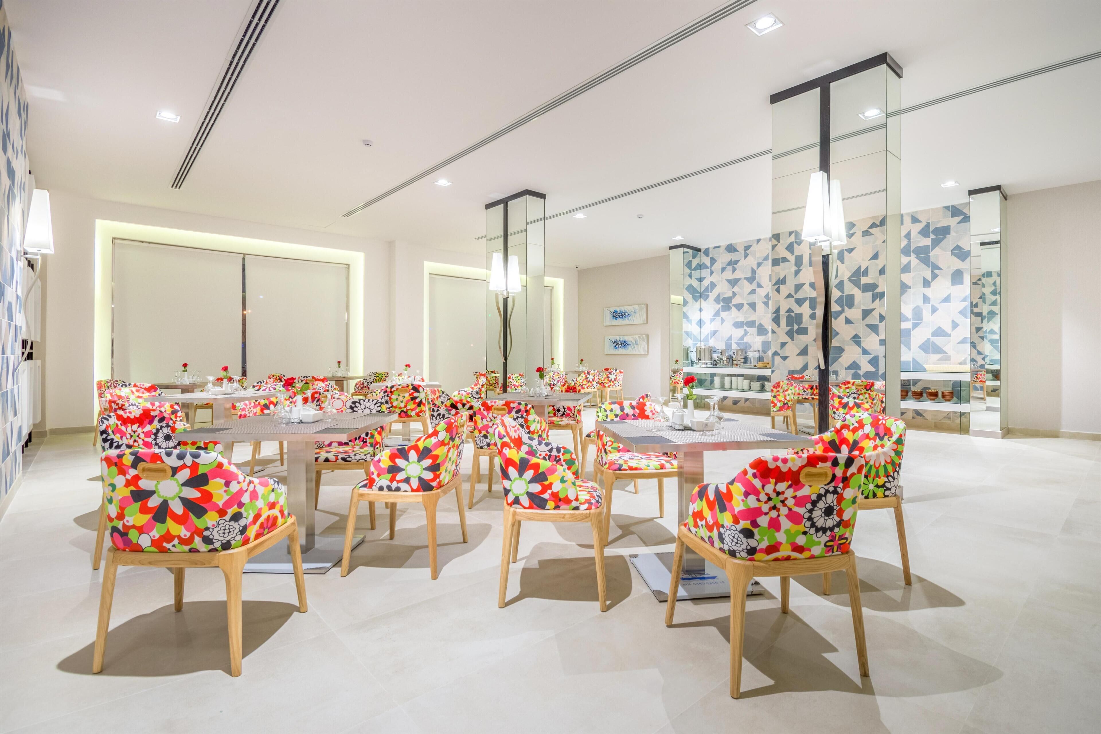 A modern dining area with multiple tables and chairs, large windows, and a decorative wall with geometric patterns.