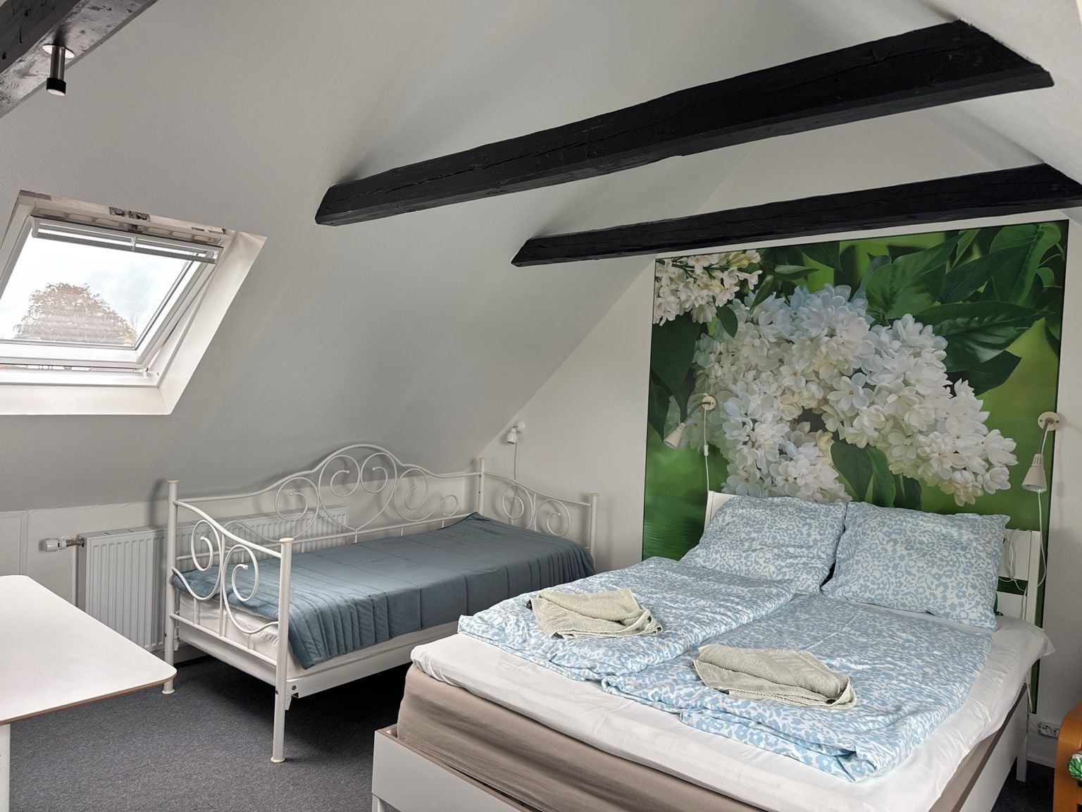 Un dormitorio con techo inclinado, tragaluces, una cama con ropa de cama estampada, una cama más pequeña y un radiador.