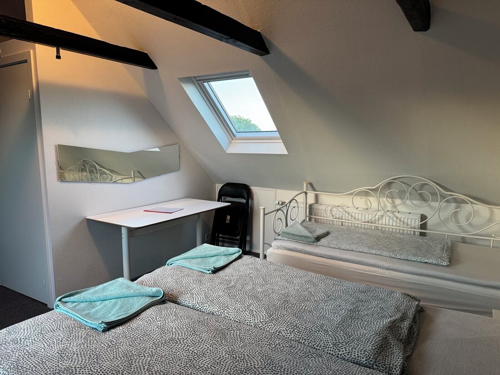 A bedroom with a sloped ceiling, a skylight, a bed with patterned bedding, a desk with a chair, and a radiator.
