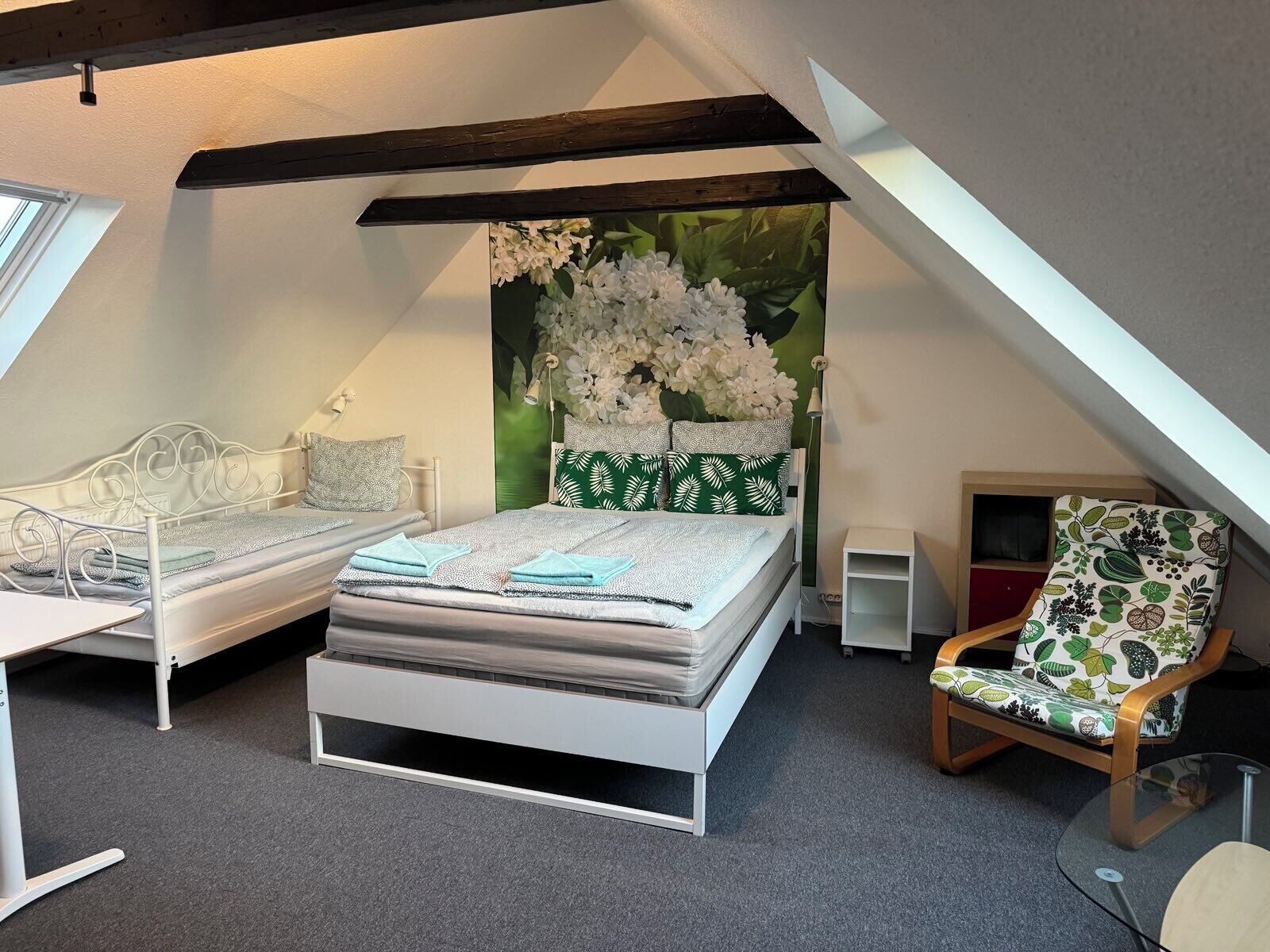 Camera mansardata con tetto inclinato, un letto con testiera a motivi verde e bianchi, una struttura in metallo bianco, un tavolino e una sedia con motivo a foglia.