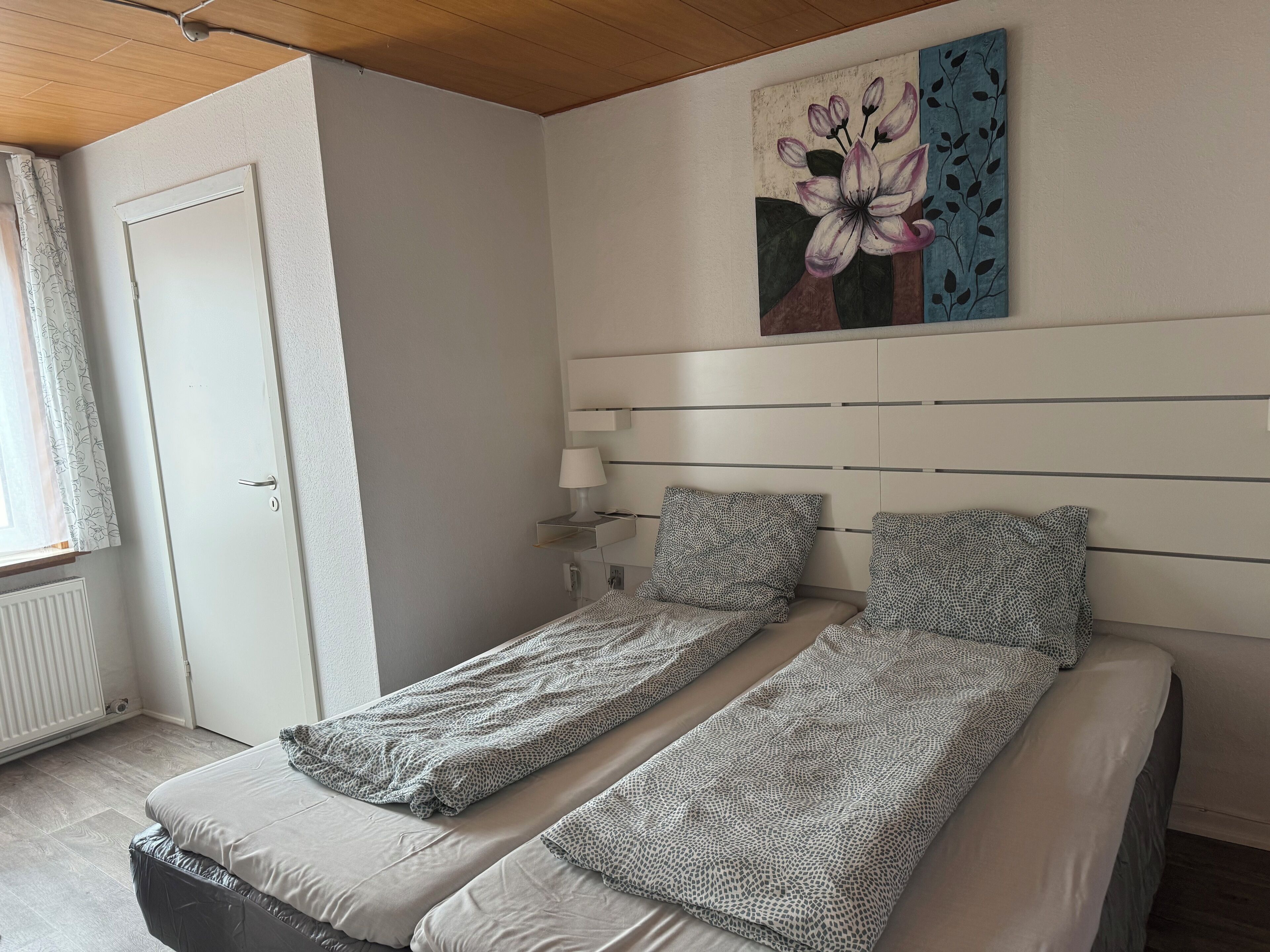 Una stanza con due letti singoli, un comodino con una lampada, un quadro appeso al muro e una finestra con tende.