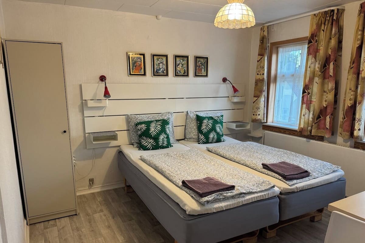 A double room with two beds, a bedside table, a lamp, a window with curtains, and a wall-mounted shelf with framed pictures.