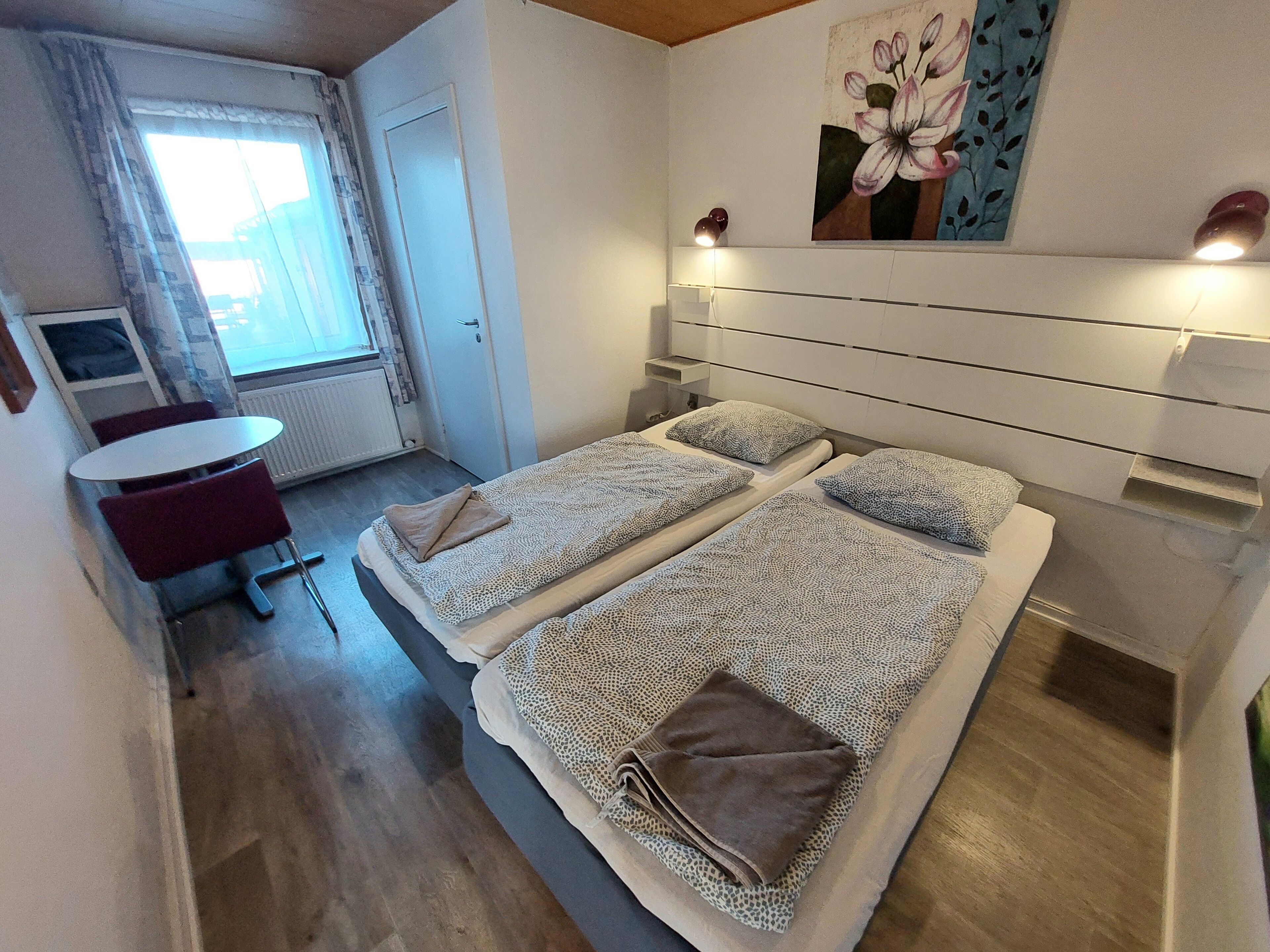 Quarto de hotel com duas camas, uma pequena mesa e uma cadeira. Há uma janela com cortinas, um radiador e um quadro na parede.