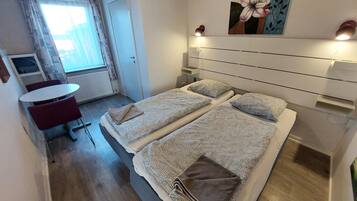 Quarto de hotel com duas camas, uma pequena mesa e uma cadeira. Há uma janela com cortinas, um radiador e um quadro na parede.