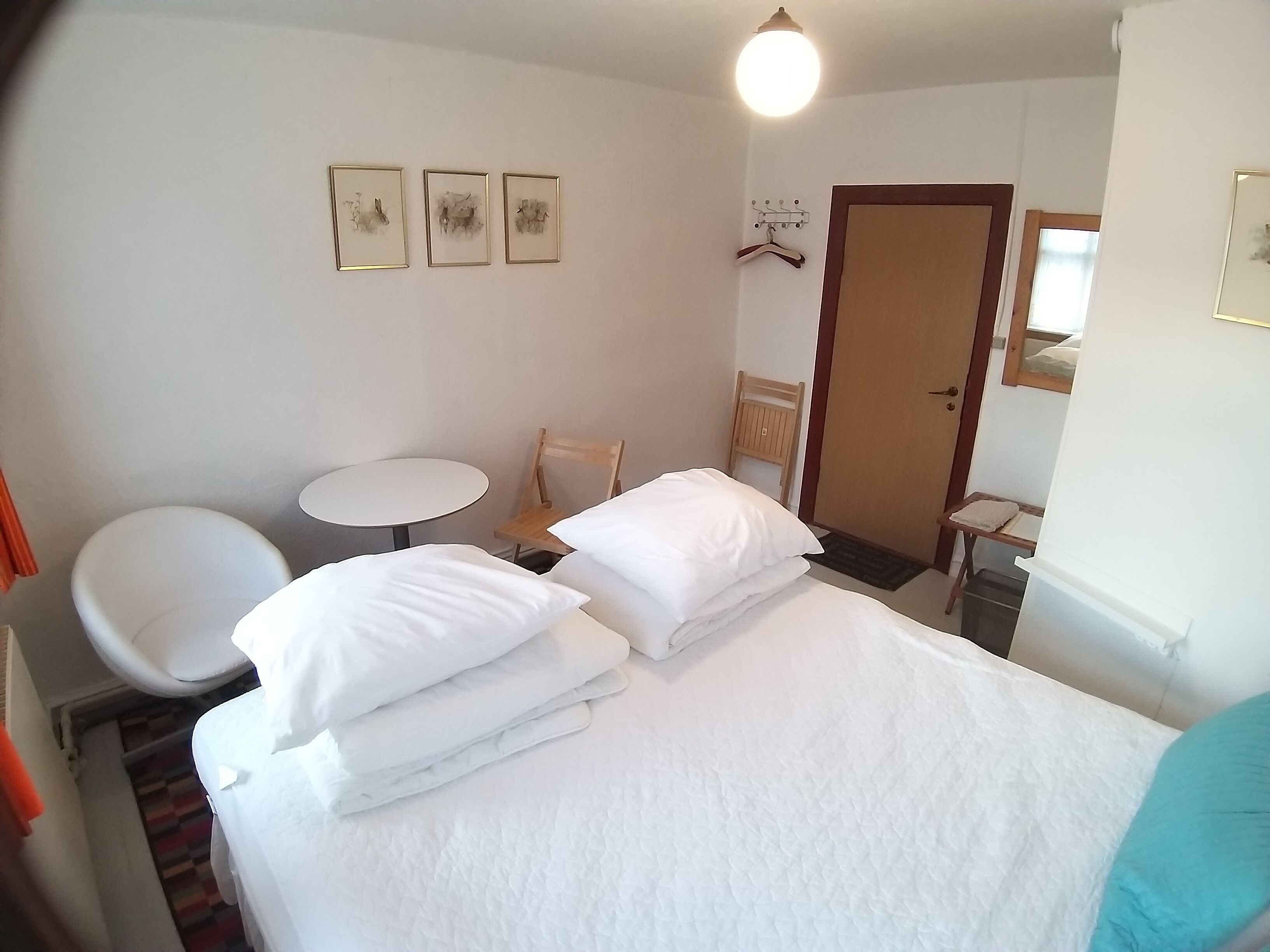 A hotel room with a bed, a chair, a small table, and a door. There are three framed pictures on the wall.
