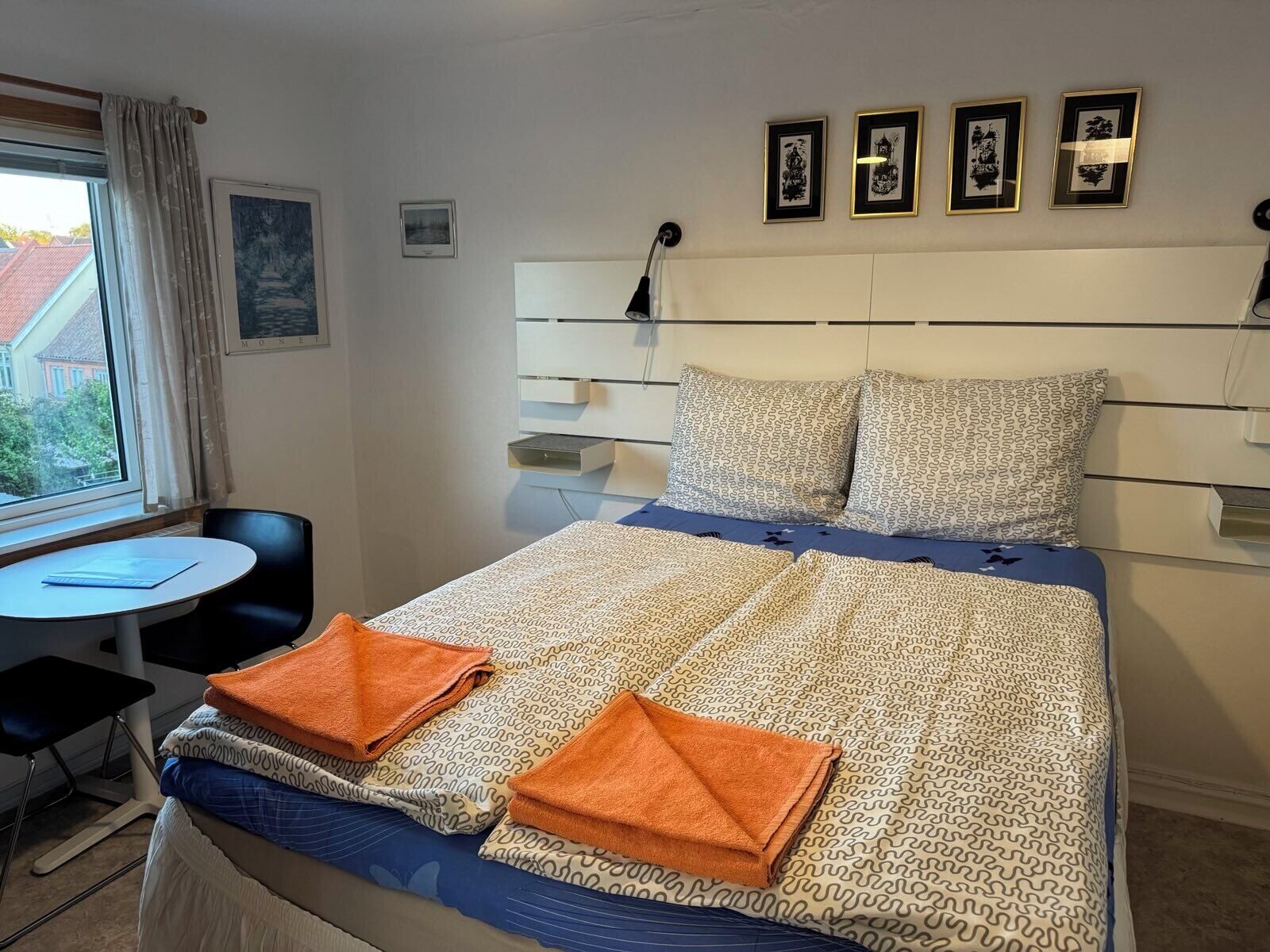 A neatly made bed with patterned bedding, a bedside table, a chair, and a window with curtains.
