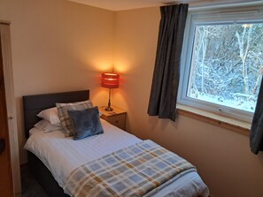 A bedroom with a bed, a bedside lamp, a window with curtains, and a view of trees outside. - Roe Deer Cottage (Beauly)