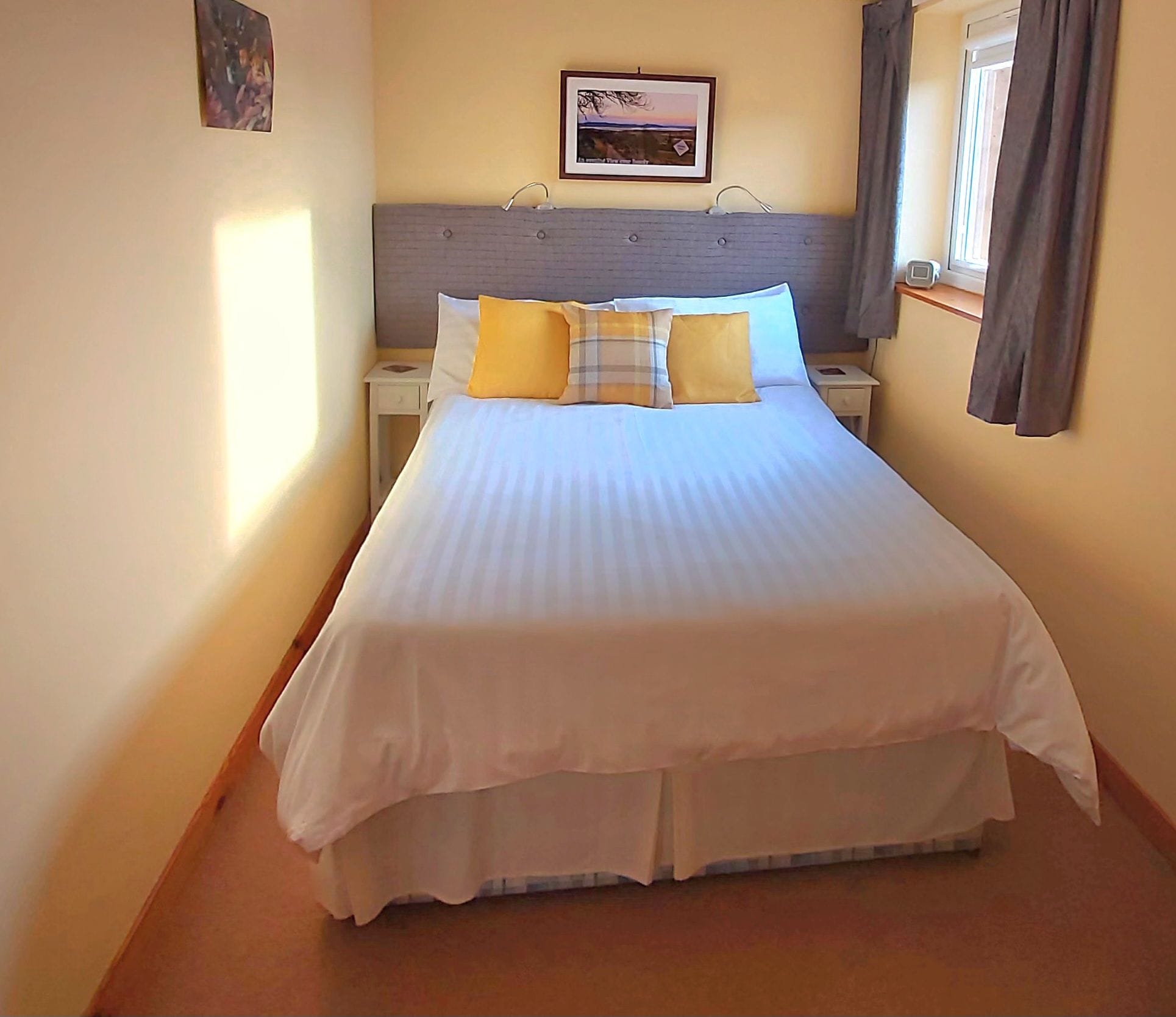 A neatly made bed with a white duvet and multiple pillows, a bedside table with a lamp, and a framed picture on the wall.