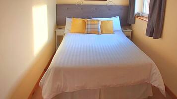 A neatly made bed with a white duvet and multiple pillows, a bedside table with a lamp, and a framed picture on the wall.