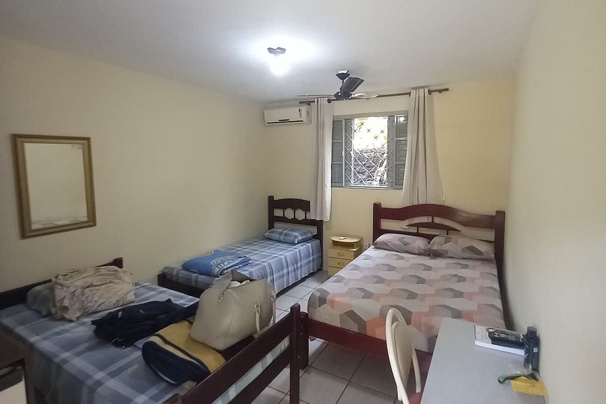 A room with three beds, a desk, and a chair. There is a window with curtains, an air conditioning unit, and a ceiling fan.