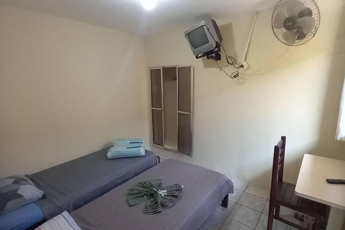 A small, simple room with two beds, a desk, a chair, a ceiling fan, and a mounted television.