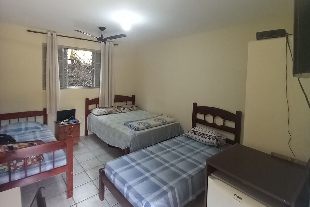 A small, simple room with two beds, a desk, a ceiling fan, a window with curtains, and a TV mounted on the wall.