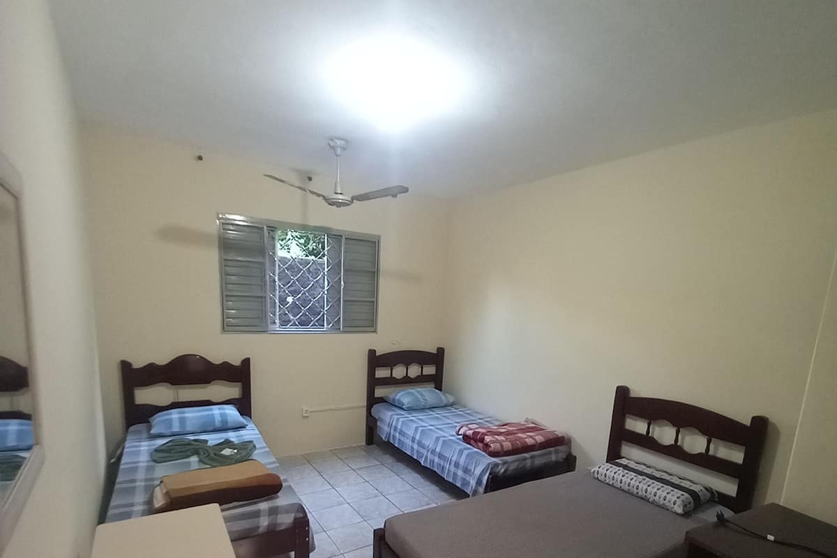 A small, well-lit room with three beds, a desk, and a ceiling fan.