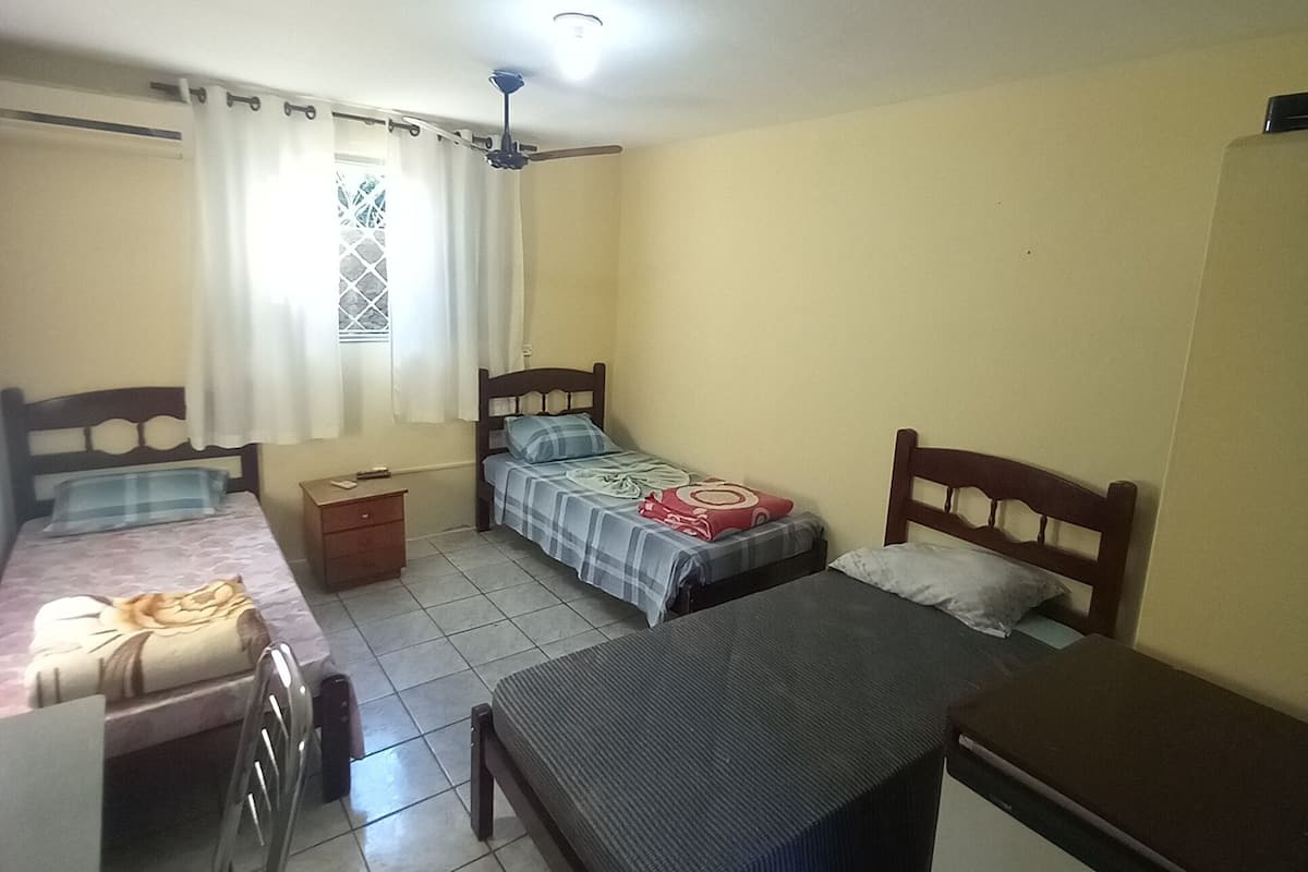 A dormitory room with three single beds, a desk, a chair, and a small cabinet.