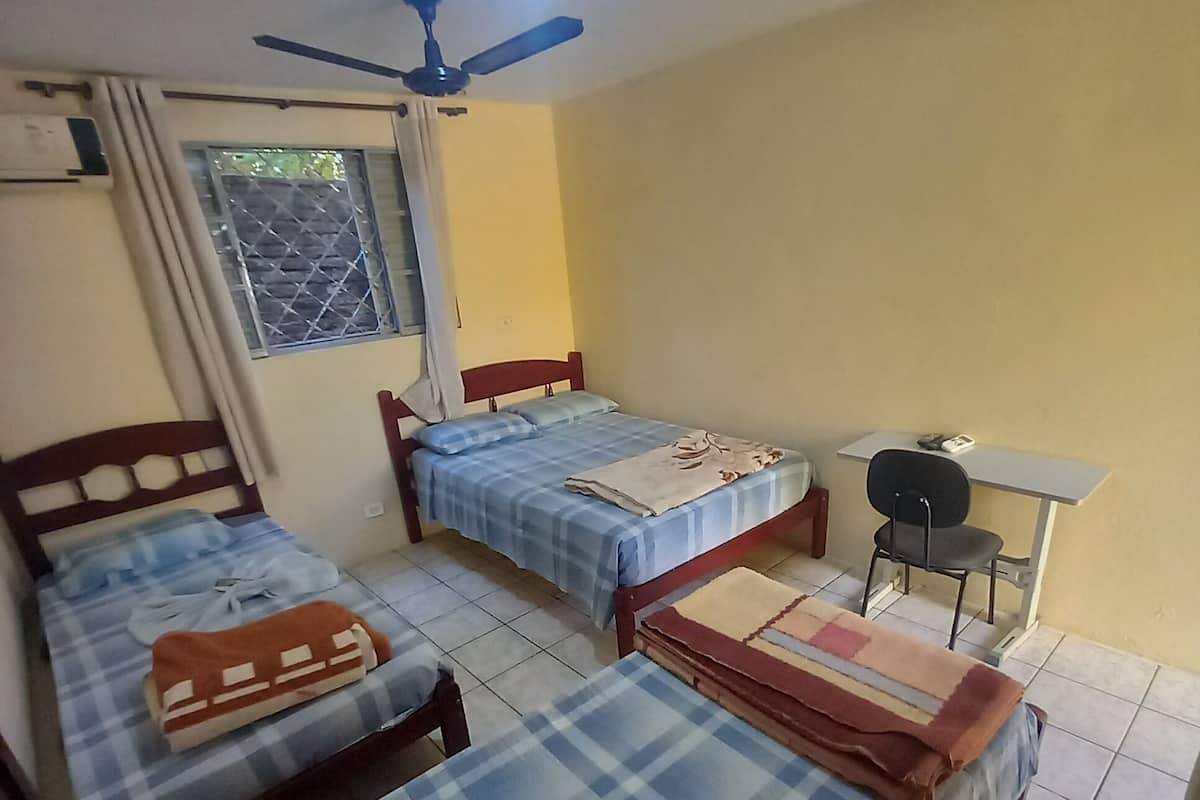 A room with two beds, a ceiling fan, a window with bars, and a small table.