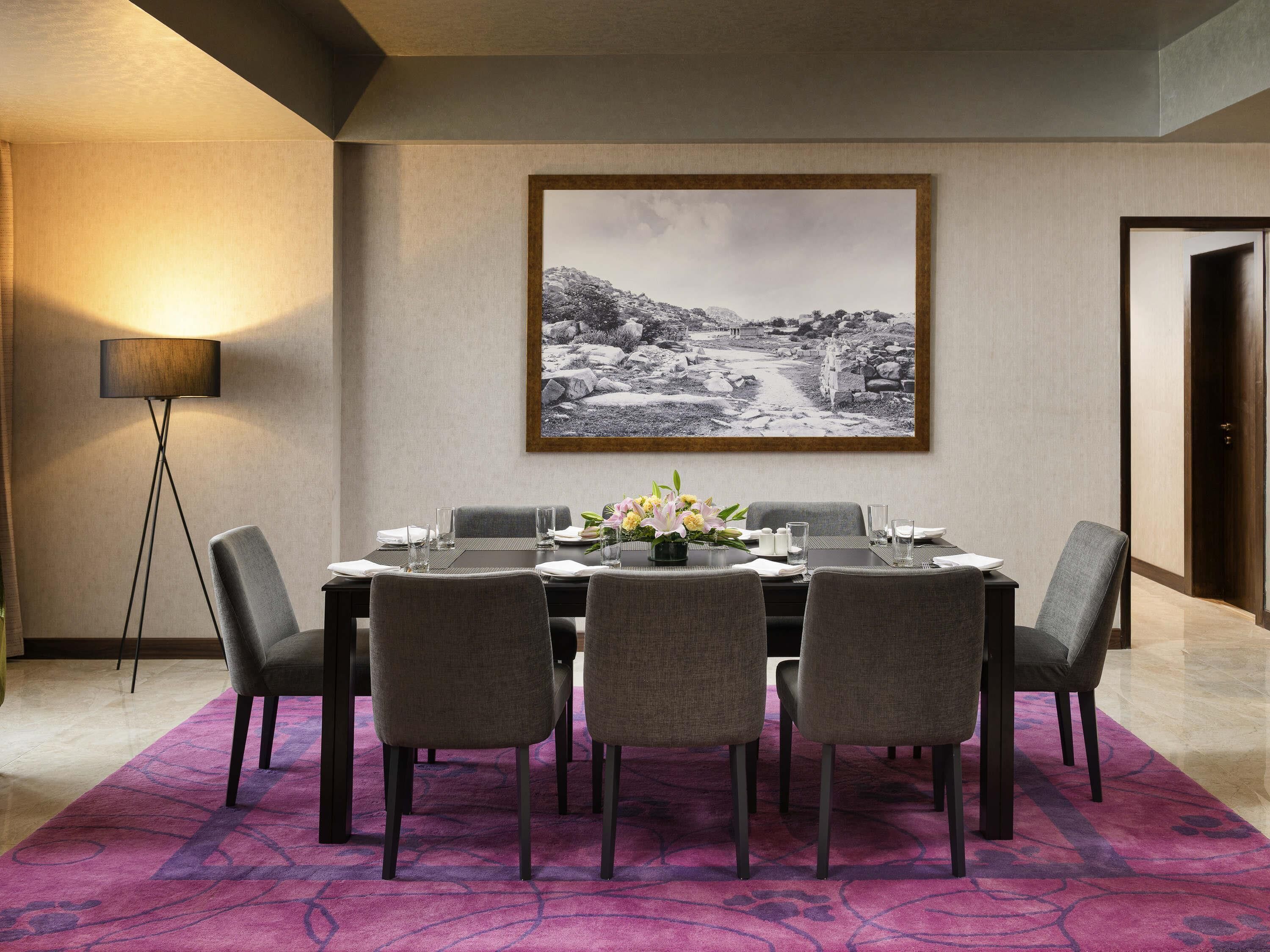 A dining area with a table set for four, a large framed picture on the wall, and a floor lamp.