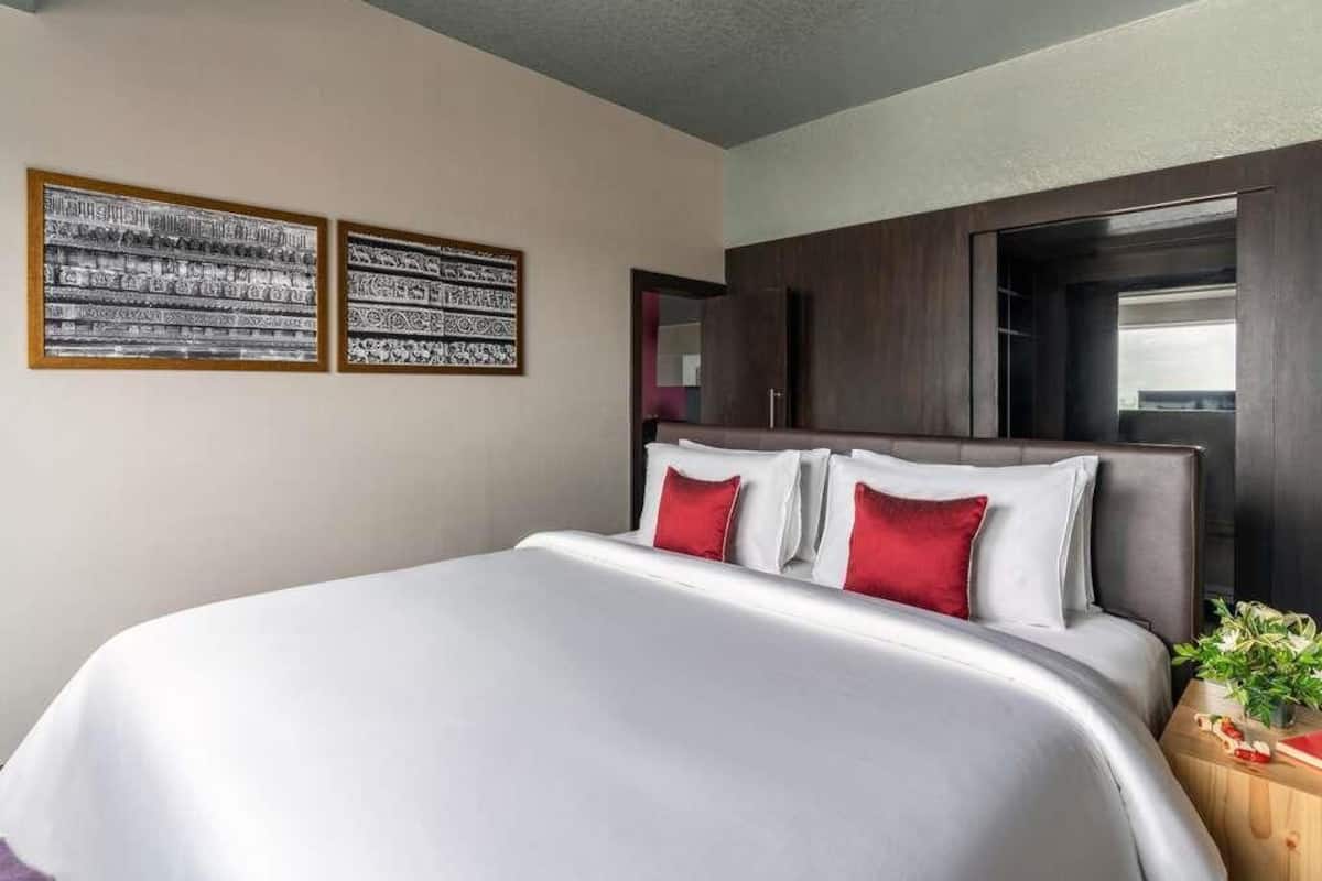 A hotel room with a large bed, two framed artworks on the wall, a wooden headboard, and a small side table with a plant.