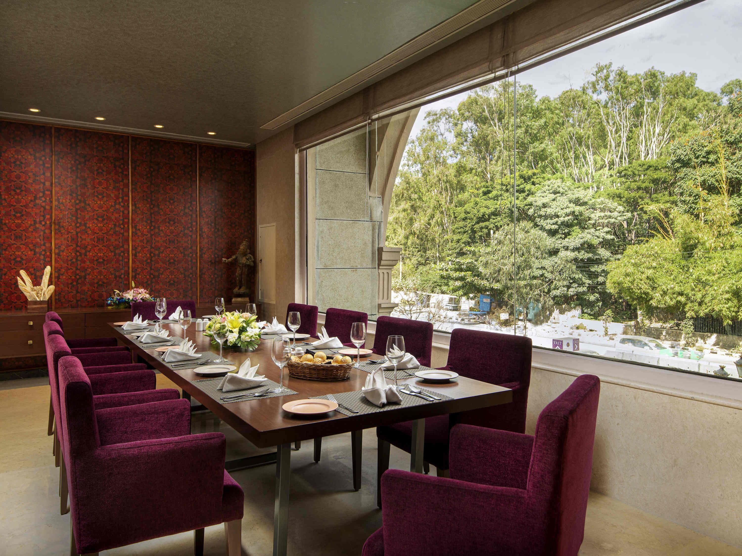 A dining area with a long table set for a meal, surrounded by chairs, with a view of trees through large windows.